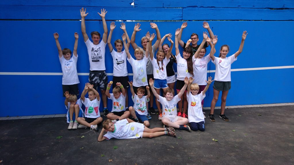 Trainer Fredi, Betreuer Sven und Hannah und 14 Kinder mit selbst bemalten Erinnerungs-T-Shirts; strecken vor der Ballwand die Arme in die Luft